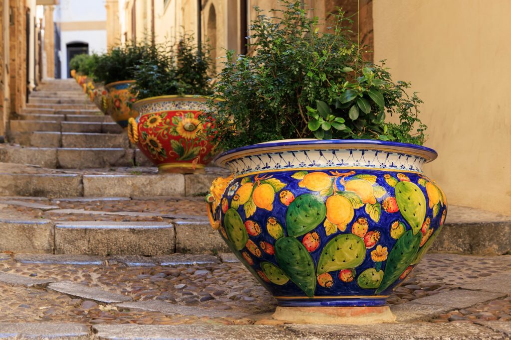 Bright Sicilian ceramic pots with plants decorate a narrow stone staircase in Cefalu, creating a vibrant and picturesque Mediterranean street scene.