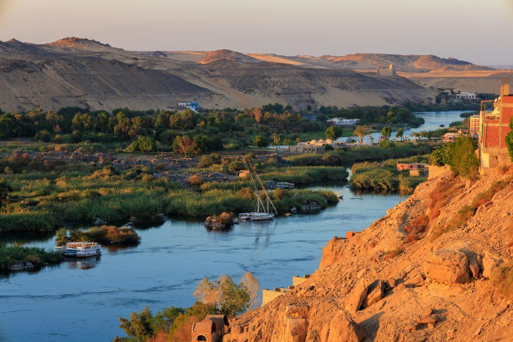 Vue du Nil serpentant entre les palmeraies, les bateaux traditionnels et les collines désertiques au coucher du soleil. Assouan, Égypte.